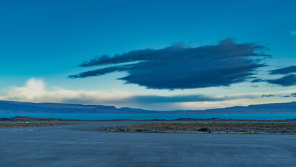 The runway of the airfield. The weather vane flutters in the wind. Low-growing grassy vegetation along the road. Turquoise glacial lake in the distance.Mountains against the sky and clouds.El Calafate