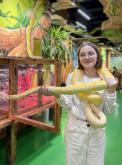 A person stands in a vibrant reptile exhibit, holding a large yellow and white snake which drapes...