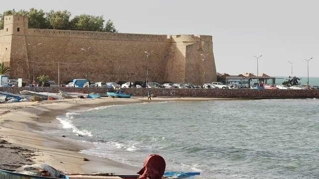 Ancient fortified citadel of Hammamet with formidable stone walls standing guard over tranquil Mediterranean seashore with old fishing boats of locals, Tunisia