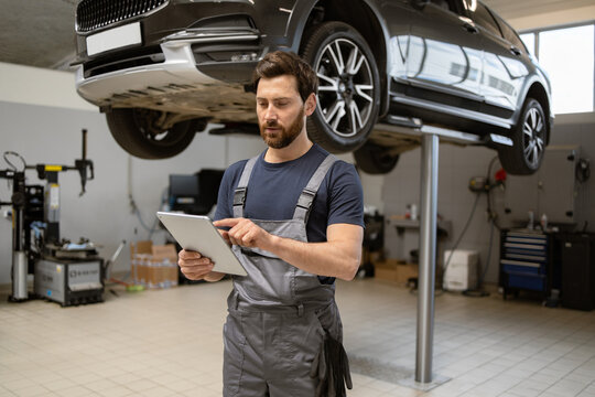 Mechanic using digital tablet in car repair shop with raised vehicle in background. Man in work uniform checking diagnostics. Auto service, modern technology in automobile maintenance.