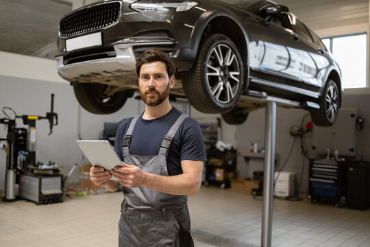Mechanic holding tablet standing in workshop with lifted car for inspection. Modern technology, automotive repair, and diagnostics concept
