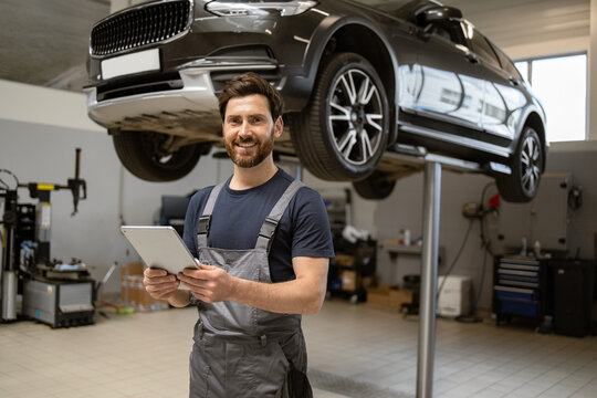 Friendly mechanic using tablet in modern car repair shop with car on lift. Professional technician checking vehicle diagnostics and maintenance. Concept of automotive industry and customer service.