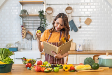 Asian young woman holds a book with online audience or simply enjoying the cooking process, health, fruit, freshness, and organic living, digital recipe platform, eating, healthy at kitchen