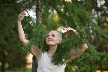  lifestyle portrait senior woman  with gray hair relaxes in summer outdoor in fir tree branches. spends time in nature.  greyhairdontcare.  Earth Day. World Environment Day. aroma therapy. 