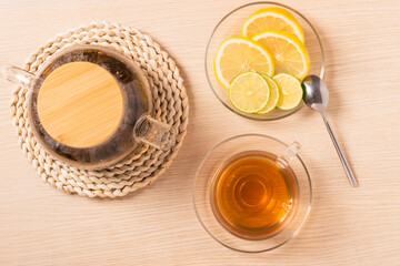 Top view of glass teapot with wooden lid, cup of tea, lemon and lime slices on wooden table