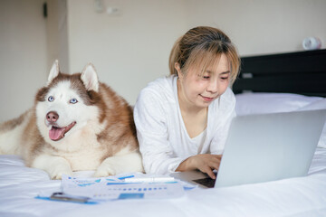 Woman using laptop while lying on bed with a husky dog ​​lying nearby © littlepigpower