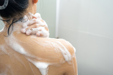 Female shoulder with gel or shampoo foam.Young woman taking shower with moisturizing soap in bathroom. Attractive girl rubbing body skin with white bath puff. Enjoying routine procedure at home.