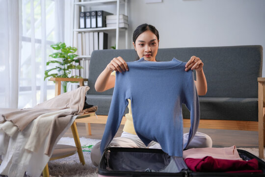 Young woman is packing for a trip, carefully folding and placing a blue sweater into her suitcase on the floor of her modern living room