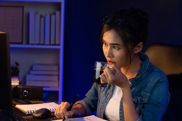 Thinking young beautiful Asian creative woman analyzing profit while drinking coffee, comparing on email sending business market report, writing on notebook for memo at neon light at night. Stratagem.