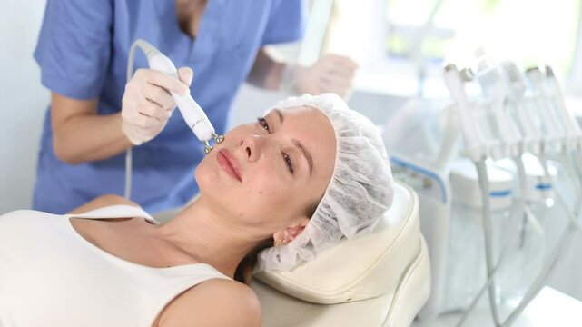 Young woman cosmetologist performs hardware facial microcurrent procedure to young female patient