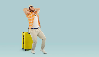 Full body photo of a funny young happy man in beach hat with yellow suitcase isolated on studio blue background. Tourist is going on summer holiday trip. Vacation and travel concept.