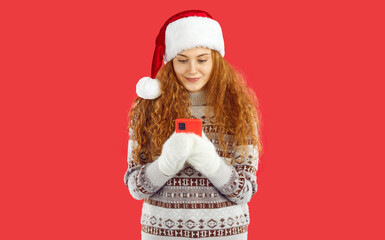 Curly pretty redhead Christmas girl in Santa hat, mittens and knitted sweater with ornament pattern is looking at smartphone on red background. Reading New Year's greetings. Winter holidays concept.