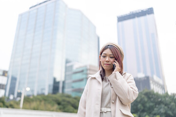 Thirty-something Hong Kong woman with reddish-brown hair talking on her smartphone, set against the backdrop of tall building cluster in Kowloon Park, Tsim Sha Tsui, Kowloon Island, Hong Kong.
