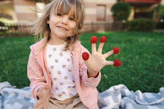 Happy girl sitting with raspberries on fingers in garden