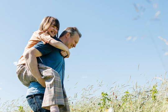 Happy father piggybacking daughter in meadow under blue sky