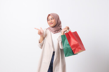 Portrait of excited Asian hijab woman in casual suit holding paper shopping bags while pointing with finger. Shopaholic girl and discount buying concept. Isolated image on white background