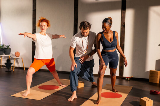 A yoga instructor guides two women in a yoga studio. The women are performing yoga poses while the instructor assists them. The studio has large windows and a calm atmosphere.