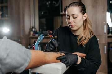Young woman professional tattoo artist in black gloves making a rose tattoo on a man's forearm.