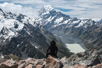 A lone hiker stands on rocky terrain, overlooking a breathtaking snow-capped mountain range with a cloudy sky. The scene includes a vast, serene landscape.