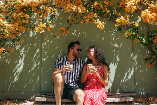 Happy couple enjoying iced drinks sitting in shade near wall