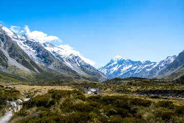 A breathtaking view of Aoraki/Mount Cook National Park in New Zealand, showcasing the snow-capped peaks, lush green valleys, and a winding hiking trail under a clear blue sky.