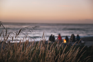A group of people gathers on the beach around a small fire at sunset, with the sky turning soft pink and orange hues, and the ocean waves nearby.