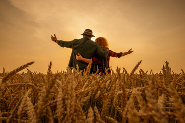 Happy man and woman are standing in their agricultural field in sunset. They are cultivating wheat and enjoying good agricultural season.