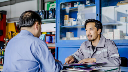 Obraz premium Professional vehicle maintenance man holding clipboard and talk with asian man customer at car service garage. Car repair and customer service concept, Discussing the cost of car repairs at the desk