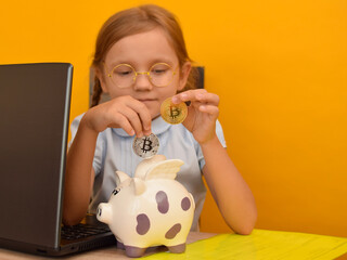 A unique child, a little cute girl sitting near a laptop puts Bitcoin coins into a piggy bank. A child learns from childhood to earn cryptocurrency. The child is a young genius, businessman.