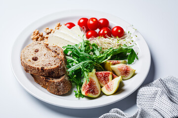 Breakfast plate with bread, tomatoes, cheese, arugula, nuts and figs on white background.