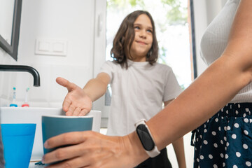 Fototapeta premium The child in the bathroom points to a rinsing cup, suggesting to use it. The mother stands nearby, observing to ensure the morning routines of brushing teeth and washing the face are done correctly.