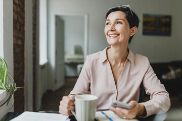 Happy beautiful mature woman in blouse with glasses on hand holding smartphone sitting at office...