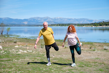 A man and woman are stretching in a field by a lake. Mature people exercising.