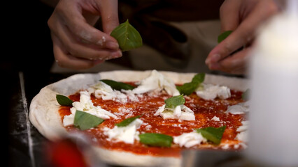 Hands meticulously add cheese and basil leaves onto pizza dough. Italian cooking