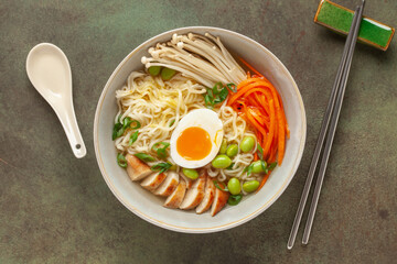 Asian ramen noodles with chicken slice, enoki mushrooms, carrots, edamame beans and egg in a  bowl. Top view