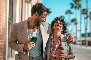 A cheerful businessman and businesswoman share a laugh on a sunny day. Clad in smart attire, he holds a smartphone while she enjoys her coffee.