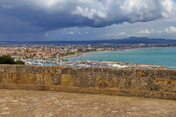 View of Palma de Mallorca from Bellver Castle featuring the marina in the foreground, notable landmarks within the cityscape, and the coastline stretching into the distance beneath a cloudy sky