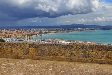 Panoramic view of Palma de Mallorca from Bellver Castle with the marina in the foreground, the cityscape including notable landmarks, and the coastline extending into the distance under a cloudy sky