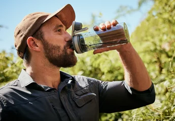 Gordijnen Trekking, man and drinking water in forest outdoor on vacation, holiday or travel on adventure. Hiking, nature or thirsty person with bottle for refreshment, health or hydration on break in woods © SneakyPeakPoints/peopleimages.com