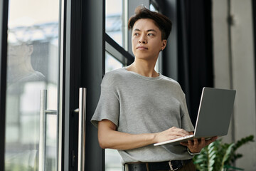 A young man stands by a window, laptop in hand, looking thoughtfully out at the city. © Bliss