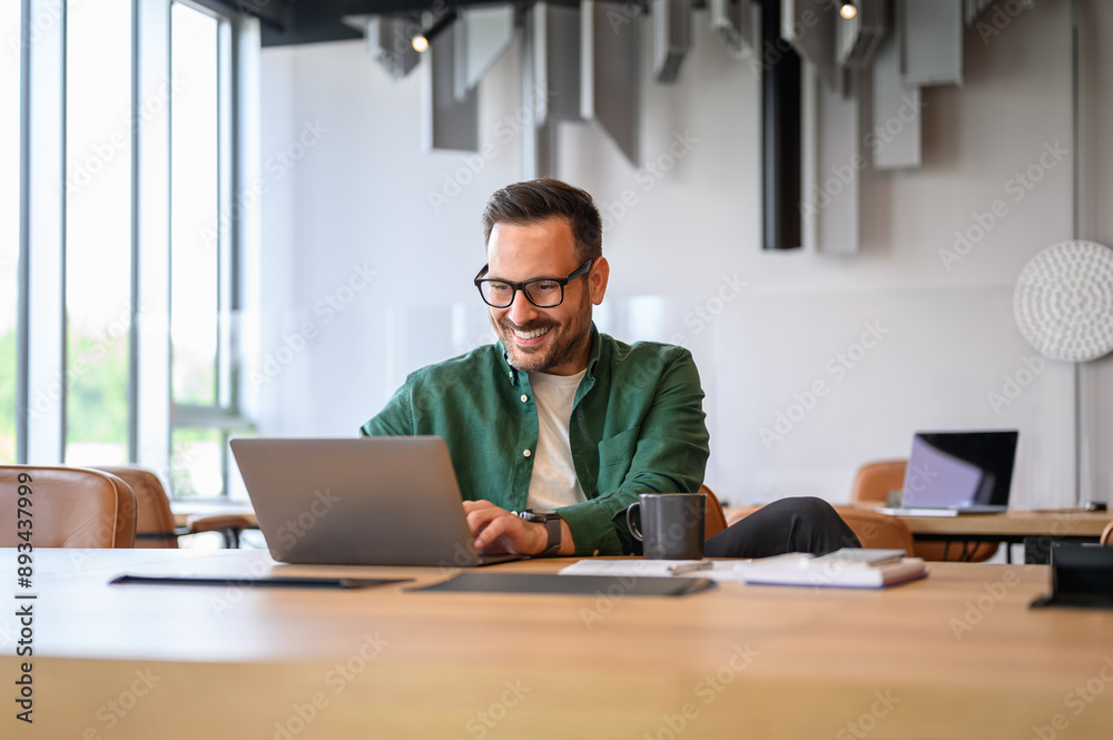 Wall mural confident handsome programmer in eyeglasses developing software over laptop at desk in modern office