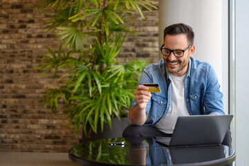 Happy successful businessman making online payment with laptop and credit card on table in office