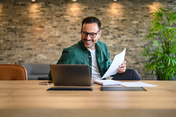 Positive male professional smiling and analyzing report over computer while working at desk in...