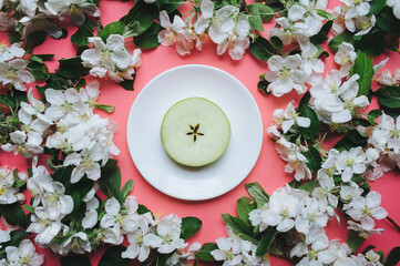 A white saucer with a round piece of apple stands on a pink background surrounded by white flowers. Close-up, top view.