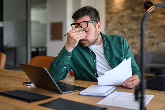 Anxious male financial advisor rubbing eyes while working over laptop and analyzing reports at desk - Powered by Adobe