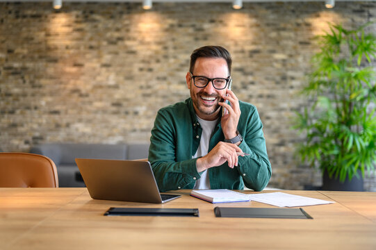 Portrait of young smiling cheerful businessman entrepreneur in casual office making phone call while working with laptop computer