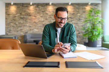 Portrait of smiling businessman reading messages over mobile phone while working over laptop at desk