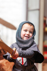 A kid is dressed in a knight costume and holds a wooden sword while playing smiling in the playroom...