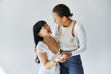 Two young women, one African American and one Asian, stand close and smile at each other, their hands clasped together.
