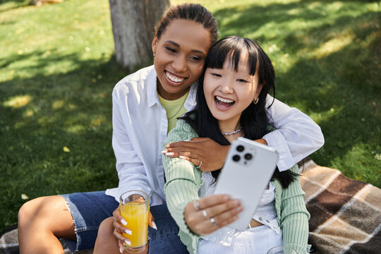 A lesbian couple, one African American and one Asian, enjoy a sunny picnic in the park while taking a selfie.